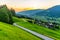 Romantic sunset in the mountains. Alpine rural curvy road, green meadows and high peaks of Austrian Alps. Pinzgau