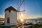 Romantic view of traditional Greek whitewashed windmill on hill. Chora town and harbor of Mykonos, Greece, low sun above