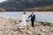 Romantic young bridal couple holding their hands on pebble riverside with forest hills as background