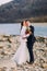 Romantic young bridal couple hugging on pebble riverside with forest hills as background