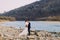 Romantic young bridal couple posing on pebble riverside with forest hills as background