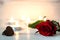 Rose lying on white a wooden surface. In the background are lit small candles. Next to the flower is two chocolates in the sha