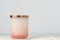 Rose pink and white ombre candle jar with shiny gold lid, sitting on a bedside table night stand. Candle is not lit. Daytime