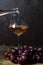 Rose wine is poured into a glass goblet from a bottle. In the foreground, a bunch of grapes is out of focus. The dark key.