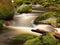 Rot trunk blocked between boulders at stream bank above bright blurred waves. Big mossy stones in clear water of river.