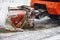 Rotating industrial brush of an orange tractor on a city street during a snowfall. Snow removal, brush machine in winter
