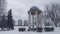 A rotunda with concrete columns stands near the walkway at the edge of the city park. Next to it are trees, and behind them is a r