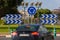 A round blue warning label. Here is a roundabout. A car in the foreground.