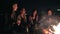 Round camera movement: Multiethnic group of young people sitting by the bonfire late at night and singing songs and