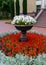 A round flowerbed with red flowers on which stands a large, brown vase with white flowers