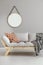 Round mirror hanging on the wall in bright living room interior with light grey sofa with cushion and blanket in real photo