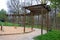Round pergola, trellises made of wooden poles around the sandpit with play elements for children. around is a path and lawns. benc