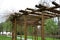 Round pergola, trellises made of wooden poles around the sandpit with play elements for children. around is a path and lawns. benc