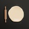 Round piece of realistic dough and wooden rolling pin lying on black table