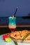 Round pizza on wooden board and Blue Hawaii cocktail on restaurant table by the lake