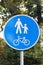 The round-shaped road sign is a pedestrian and bicycle zone against a background of green foliage. White people sign and bicycle