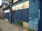 A row of abandoned stores with boarded up shop fronts with crumbling facades and peeling blue paint in an urban street