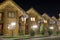 Row of new two-storied ecological wooden comfortable decorated cottages hotels on clean illuminated street under dark night sky.