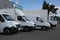 Row of white delivery and service van, trucks and cars in front of a factory and warehouse