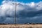 Row of wooden electricity poles with three wires and ceramic isolators in field. Heavy dark blue clouds before thunderstorm, brown