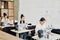 Row of young seamstresses sitting by desks and using electric sewing machines