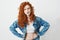 Rude young girl with red curly hair looking at camera brutally with arms akimbo over white background.