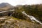 Rugged Welsh landscape, with a road and bridge over fast flowing river, rugged rocks, fir trees and low cloud ov er the mountains