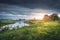 Rural landscape of river nature with cloudy sky at sunrise. Scenic nature in the morning. Green meadow near river. Bad weather in