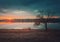 Rural landscape with sunset reflecting over the calm lake water, a bare willow tree on the dry grass meadow and a wanderer man