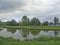 Rural pond with green grass and trees under a cloudy sky