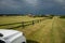 Rural view, stormy clouds and green field with white car and wooden corral like fence.