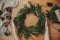 Rustic Christmas wreath, flat lay. Hands holding christmas wreath with fir branches, berries, pine cones, and thread, scissors on