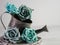 Rustic metal watering can with a gray wooden wreath and 4 teal and blue paper rose flower blooms and a soft gray background.