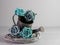 Rustic metal watering can sitting on a gray wooden wreath coiled up with blue and teal paper rose flower blooms on a sandstone