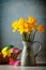 Rustic styled still life of bunch of yellow daffodils in vintage water jug with Easter decorations. Selective focus, textured