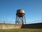 Rusting water tower stands beyond the wall and bard wire fence