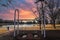 A rusty white metal swing set in the park in front of a still green lake surrounded by yellow winter grass and bare winter trees