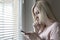 Sad apathetic lonely woman standing near the window and texting messages in smartphone at home or hotel, divorce, depression and