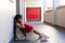 Sad biracial schoolgirl with school bag sitting at wall at elementary school corridor, copy space