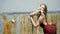 A sad blonde with long hair in a red dress holds onto wooden posts under the gusts of wind, in the middle of the estuary