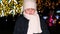 A sad boy in a white hat and scarf looks down standing near the Christmas tree in the evening.