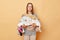 Sad brown haired Caucasian woman standing with multicolored laundry cluttered with clothes collects clothing for recycling