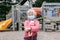 Sad Caucasian girl in face mask with baby toy on closed playground outdoor. Kids play area locked with yellow caution tape in