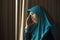 Sad and depressed Muslim woman in Islam traditional Hijab head scarf at home window feeling unwell suffering depression crisis and