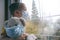 Sad illness child on home quarantine. Boy and his teddy bear both in protective medical masks sits on windowsill and looks out