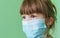 A sad little girl in a medical mask on a green background. Disposable medical mask on the child`s face.A child in a protective med