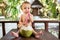 A sad little one year old child sits on a wooden table and drinks coconut milk from fresh green coconut through a straw