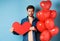 Sad man looking heartbroken and lonely, holding paper red heart and standing near balloons over blue background