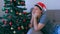 Sad man in red cap is boring sitting on couch near Christmas tree at home.