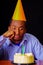 Sad man wearing blue shirt and hat sitting by table with cake in front, single candle burning, looking bored depressed